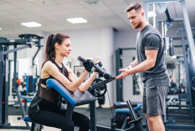 Esmer fitness kızı spor salonunda halter diskiyle antrenman yapıyor. Antrenör daha iyi sonuçlar elde etmeye yardımcı olur.
