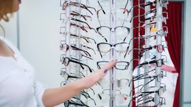 漂亮的年轻女子正在光店里挑选新眼镜 美丽的女士选择眼镜站在光学仪器前 — 图库视频影像