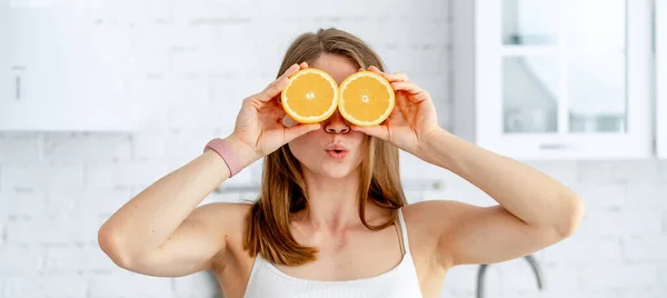 若い魅力的な女性は台所の背景の彼女の美しい顔の近くにオレンジの2つの半分を保持する 健康食品のコンセプト — ストック写真