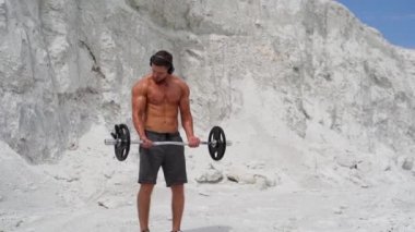 Beyaz dağ arka planında kaslı vücut geliştirici halter kaldırıyor. Kulaklıklı güçlü genç bir sporcu dışarıda antrenman yapıyor. Yavaş çekim.
