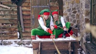 Dışarıda tahta kızakta oturan yeşil kostümlü iki elf. Mutlu elfler kışın evin arka planında eski peri kızağında birlikte otururlar. Noel zamanı.