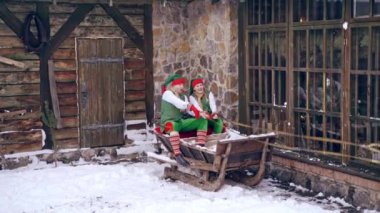 Neşeli elfler kışın dışarıda Noel 'de birlikte vakit geçiriyorlar. Noel Baba 'nın kızağında oturan peri karakterleri ahşap bina arkasında.