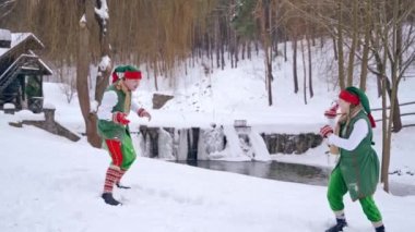 Yeşil kostümlü iki komik elf kış arka planında mutlu bir şekilde oynuyorlar. Bir cüce nehrin yanında beyaz kara düşüyor. Mutlu Noeller..