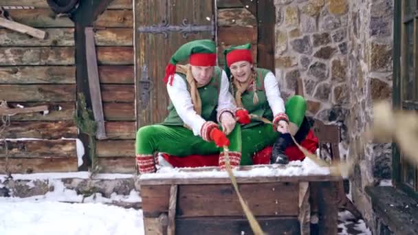 緑の衣装を木製のそりの屋外に座って2つのエルフ 幸せの葉一緒に古い妖精のそりで冬には家の背景に座っている クリスマスタイム — ストック動画