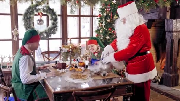 圣诞老人身穿红色服装 白胡子 把热水倒入杯子里 老态龙钟的精灵们坐在圣诞装饰背景下的节日桌旁 — 图库视频影像