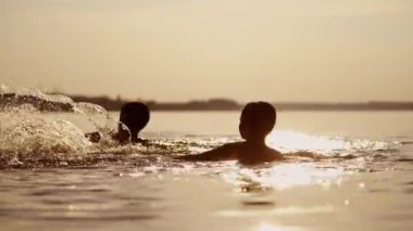 Küçük çocuklar suda oynuyor ve su sıçratıyor; çocuklar gölde veya nehirde yüzüyor ve damla damla ile eğleniyorlar; iki kardeş gün batımında rollick yapıyorlar. Monokrom video. Yan görünüm.