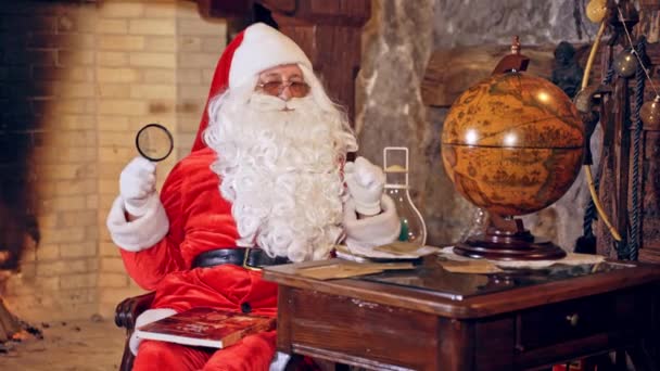 サンタクロースはテーブルに特別に装飾された部屋に座っていて 封筒を手に持っています 背景にある饗宴暖炉 クリスマスの精神の概念はビデオを閉じます クリスマス 新年が近づいています — ストック動画