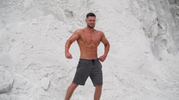 屋外で訓練された体を持つアスレチックマン 筋肉スポーツマン無気力なショートパンツでカメラに白い岩の背景 — ストック動画