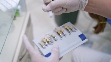 Dişçinin elinde yapay diş var. Beyaz steril eldivenli stomatolog sentetik diş rengini seçiyor. Diş implantlarının faydaları.
