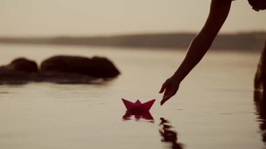 Kadının eli nehirde kırmızı ve yeşil kağıt gemileri fırlatıyor. Origami Kağıt Tekneleri gölde yüzüyor. Güzel Dalgalar Dalgalanma Arkaplanı. Seyahat ve doğa kavramı. Filtrelenmiş video.