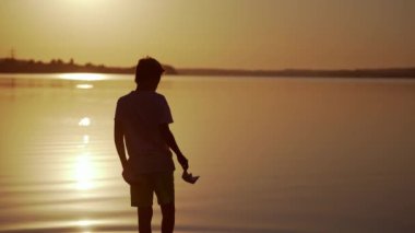 Kırmızı ve yeşil kağıtlı çocuk nehirde yürüyor. Origami Kağıt Tekneleri çocuğun elinde. Güzel Dalgalar Dalgalanma Arkaplanı. Seyahat ve doğa kavramı. Gün batımında video.