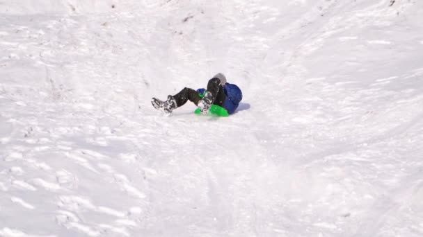 少年は雪の丘の上で眠っていた 子供は冬にプラスチック製のそりに乗ってスライドを滑り降りる 幸せな子供時代 スローモーション — ストック動画