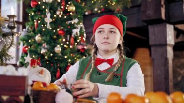 Dişi elf elinde metal bir bardakla Noel masasında oturuyor. Yeşil kostümlü kız gülüyor ve konuşuyor. Yeni yıl ve Noel konsepti.