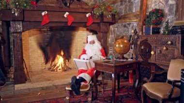 Noel Baba özel dekore edilmiş bir odada oturuyor ve kitaba bakıyor. Noel ve Yeni Yıl konsepti. Mucize zamanı. Arka planda şömine.