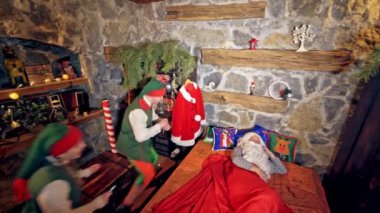 Noel Baba yatağında özel dekore edilmiş bir odada uyuyor. Elin yanağın altında. Elfler çan çalıyor ve yüzüne yakın el sallıyorlar. Noel ruhu konsepti videoyu kapat. Yukarıdan video.