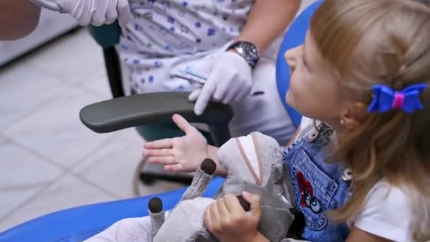 小児科の椅子の小さな子供 閉じます 歯医者のキャビネットでおもちゃでかわいい女の子 勇気のある赤ん坊は彼女の問題を正統化する ビデオを閉じます — ストック動画