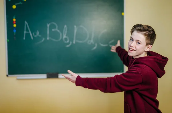 Student of higer school is standing in dark hoodie near the green blackboard in the classroom. Teenage boy is writing alphabet on the board. School education concept