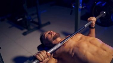 Gömleksiz genç vücut geliştirici göğüs için göğüs presi yapıyor. Güçlü karın kasları. Mavi ışık filtresi. Yan taraftan video.