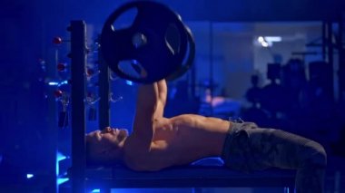 Vücutları mükemmel olan vahşi bir sporcu spor salonunda ağırlık kaldırıyor. Mavi ışık filtresi. Yan taraftan video.