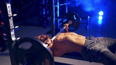 Sporcu içeride antrenman yapıyor. Genç sporcu tahtada yatıyor ve modern spor salonunda ağır halterleri kaldırıyor. Mavi ışık süzgeci.