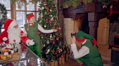 Noel ağacının yanında mutlu elf ve Noel Baba. Elf dekore edilmiş odada mutlu bir peri karakterinin fotoğrafını çekiyor..