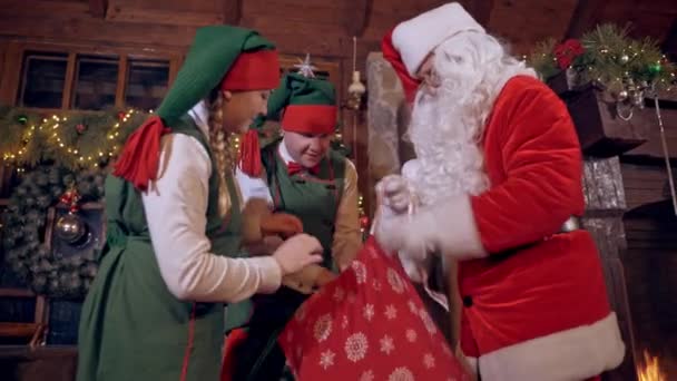 圣诞老人拿着装满玩具的红包仙女精灵在圣诞老人住所的壁炉前帮助圣诞老人 — 图库视频影像