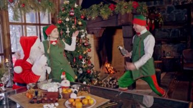Komik elf Noel 'de fotoğraf çekiyor. Noel Baba ve dişi elf Noel ağacının yanında. Retro kamera elf 'in elinde..