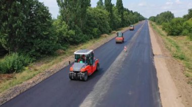 Otobanda çalışan yol silindirleri. Yeşil doğanın ortasında yolda asfaltı dümdüz eden kamyonlar. Asfalt yol inşaatı. Hava görüntüsü. Hareket kamerası geri.