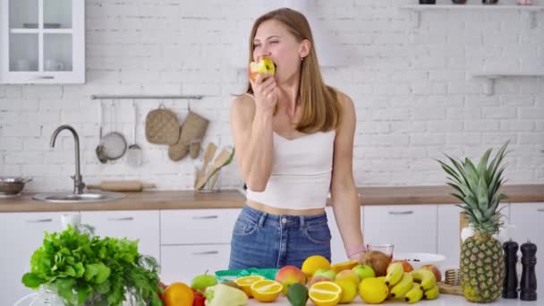 减肥的概念 年轻女人在厨房里吃水果 漂亮的牛仔裤模特站在满是有机维生素的桌旁 — 图库视频影像