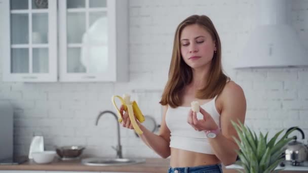 きれいな女性はバナナを食べるのが好きだ カジュアルな服装の若い女性はキッチンで新鮮な果物を食べます 健康食品のコンセプト — ストック動画