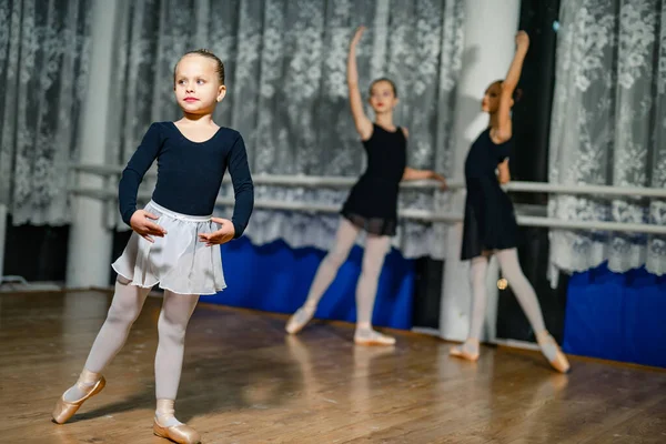 Little ballerina standing in pose with her hands on a blur background with two ballerinas. Girls practicing during the lesson in a dancing studio. Close-up
