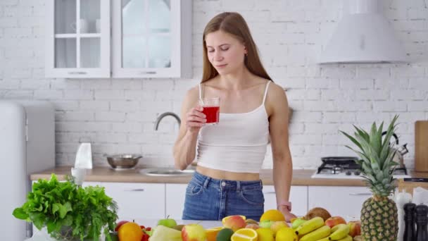 年轻女人喝新鲜果汁 一位漂亮的女士站在餐桌旁边 喝着一杯美味的果汁 在现代厨房里享用着有机水果 减肥的概念 — 图库视频影像