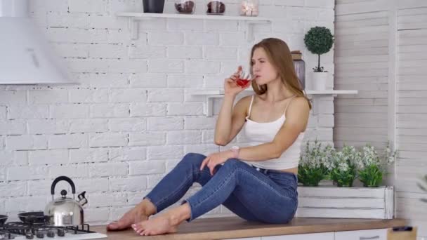 美人は家でジュースを飲む 若い女性のサイドビューでジーンズでキッチンテーブルの上に座って新鮮なジュースを飲む — ストック動画