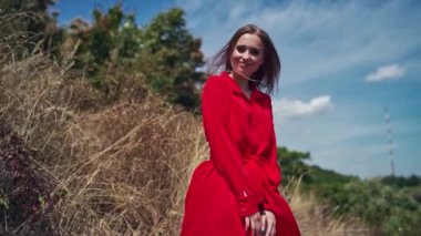 Yazın doğa arasında güzel bir model. Kameraya poz veren kırmızı elbiseli çekici bir kadın. Genç bir kadın nehir kenarındaki tepede yürüyor. Arkasını dönüyor ve gülümsüyor..