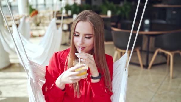 カフェで赤いドレスの魅力的な女性 若い美人さんが白いスイングでジュースを飲んでいます 美しい女性の肖像画をリラックス スローモーション — ストック動画