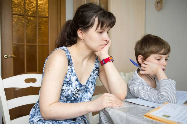 A first-grade boy with regrown hair is doing homework in a notebook with his mother.