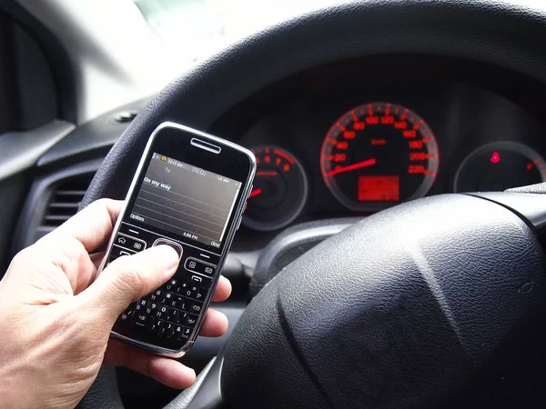 Hand holding a cellphone in front of a steering wheel of a car — Stock Photo, Image
