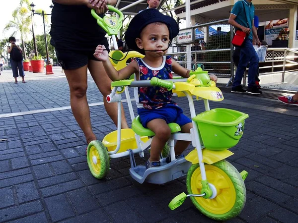 Um menino é empurrado por sua mãe em seu triciclo colorido kiddie . — Fotografia de Stock