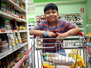 O bir markette alışveriş sepeti iter gibi genç bir çocuk kameraya gülümsüyor.