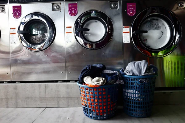 Vêtements sales dans un panier à linge devant des rangées de machines à laver à l'intérieur d'une laverie automatique . — Photo
