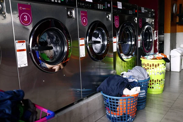 Vêtements sales dans un panier à linge devant des rangées de machines à laver à l'intérieur d'une laverie automatique . — Photo