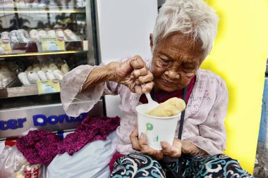 Buruşuk derili yaşlı Filipinli kadın kaldırımda otururken bir kase lapa yiyor.