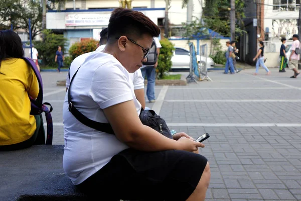 Filipino man sits on a park bench to relax and use his smartphone — Stok fotoğraf
