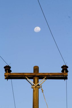 Bir elektrik direğinin, ayın ve açık mavi gökyüzünün fotoğrafı.