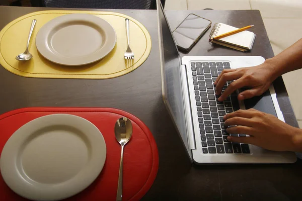 Photo of laptop computer with hands typing on it, smartphone, notebook, pencil, plates and utensils on a dining table.