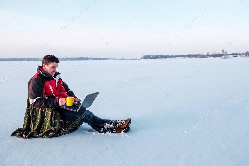 A freelancer man in warm jacket works with a laptop on the surface of a frozen lake.