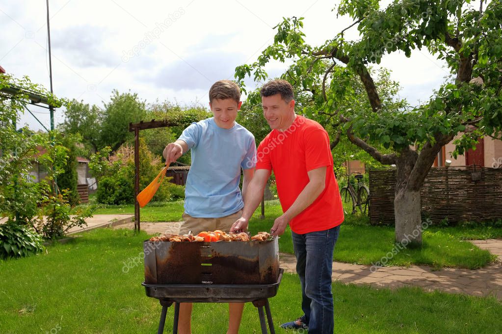 Attractive white father and son are smiling and grilling kebabs outdoors near their home