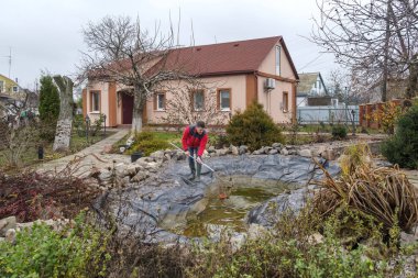 Beyaz, orta yaşlı bir adam bir bahçe havuzunu balçık, su bitkileri, düşen yapraklar ve balık ağlarıyla temizliyor..