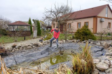 Beyaz, orta yaşlı bir adam bir bahçe havuzunu balçık, su bitkileri, düşen yapraklar ve balık ağlarıyla temizliyor.