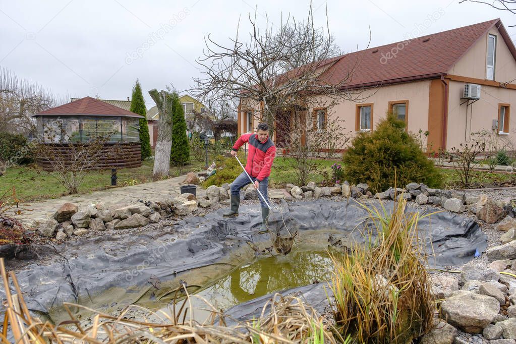 White middle-aged man cleans a garden pond with landing net from slime, water plants, falling leaves and catches fish